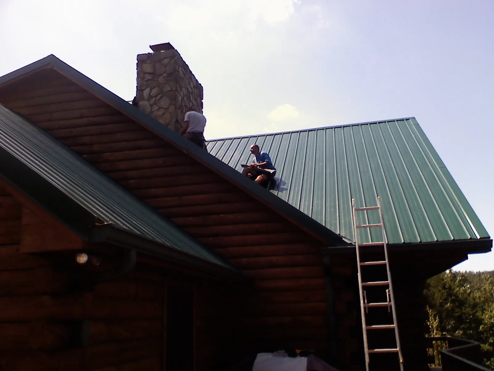 Roofers installing green metal panels beside a stone chimney on a log home in Pierce